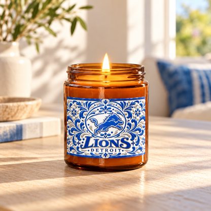 An amber glass jar candle with a lit flame sits on a wooden table in a sunlit room. The jar has a decorative blue and white label reading 'Lions Detroit' with a lion graphic.