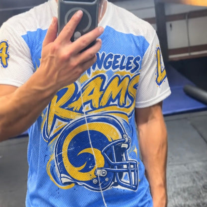 A man taking a mirror selfie in a gym wearing a Los Angeles Rams athletic t-shirt. The shirt is light blue and white with a large yellow and blue Rams helmet graphic on the front.