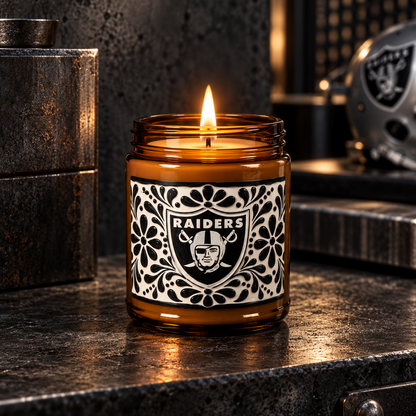 A lit amber glass jar candle with a black-and-white Raiders logo label, set on a dark metallic surface in a moody, industrial-style setting.
