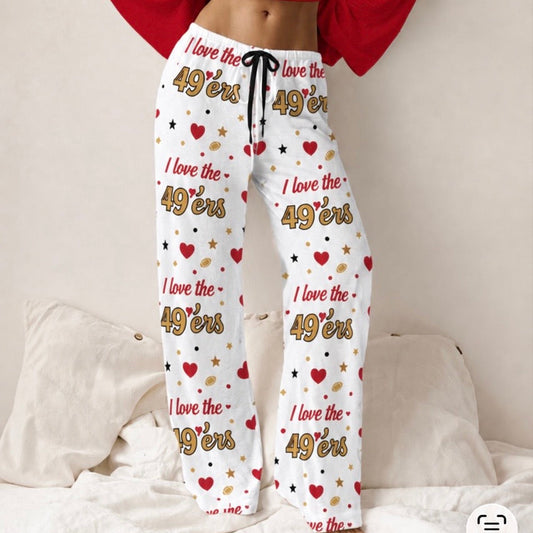 White lounge pants with a 'I love the 49ers' print featuring red hearts, gold stars, and black text, modeled on a person lying on a bed with white pillows.