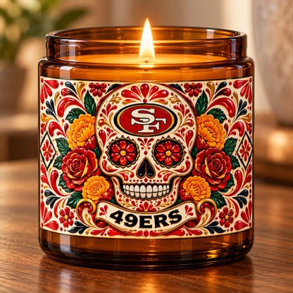 An amber glass jar candle with a lit flame, featuring a colorful Day of the Dead sugar skull label with the San Francisco 49ers logo and flowers, set on a wooden surface.