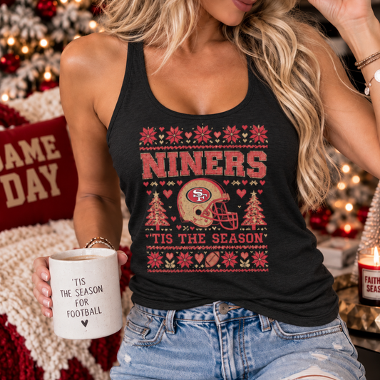 A woman wearing a black tank top with a red and gold San Francisco 49ers holiday graphic that reads 'Tis the Season for Football', paired with a matching red beanie and denim shorts, holding a mug in a cozy, decorated living room setting.