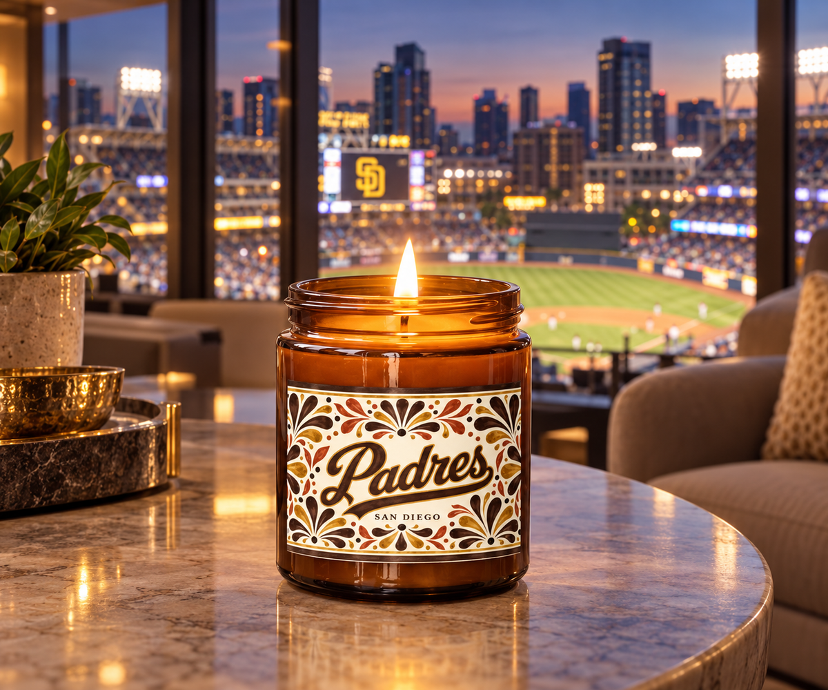 A lit amber glass jar candle with a decorative label reading 'Padres San Diego' sits on a marble table in a modern room with a large window overlooking a baseball stadium at dusk.