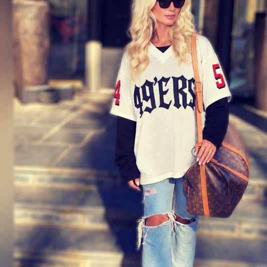 A woman walking outdoors wearing a white 49ers jersey sweatshirt layered over a black long-sleeve shirt, distressed light blue jeans, tan boots, and carrying a brown monogram shoulder bag.