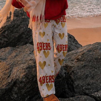 A person standing on rocks by the ocean at sunset, wearing a red cap with a white logo, a red sweatshirt, a cream scarf, and cream pants with red and gold 49ers lettering and heart patterns.