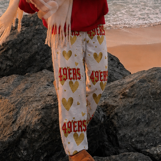 A person standing on rocks by the ocean at sunset, wearing a red cap with a white logo, a red sweatshirt, a cream scarf, and cream pants with red and gold 49ers lettering and heart patterns.
