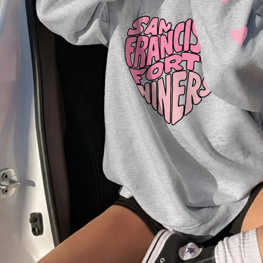 A woman sitting in a car wearing an oversized light gray sweatshirt with a pink graphic that reads 'San Francisco Forty niners' and small pink hearts on the sleeves.