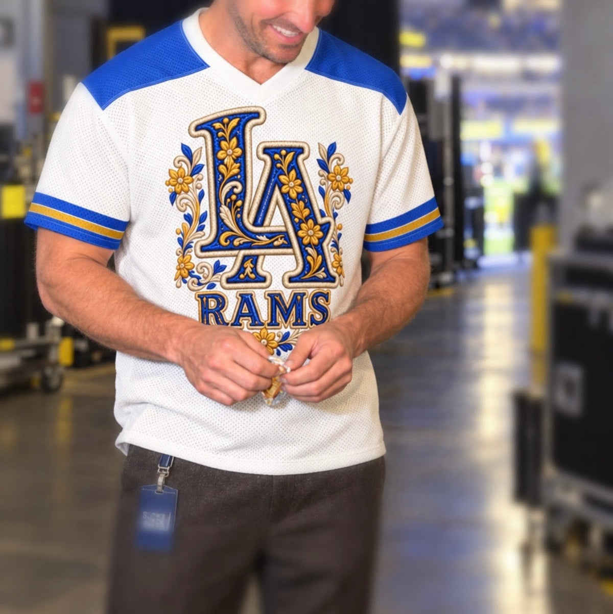 A man wearing a white Los Angeles Rams athletic t-shirt with blue and gold details, standing in a large indoor arena or event space.